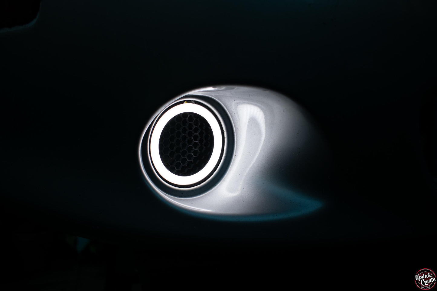 LED Halo light mounted to a custom 3D printed housing installed onto an OEM silver Mazda Mx5 NB bumper. The light is turned on, emitting a white colour. The item is pictured at night to show its brightness in the dark.