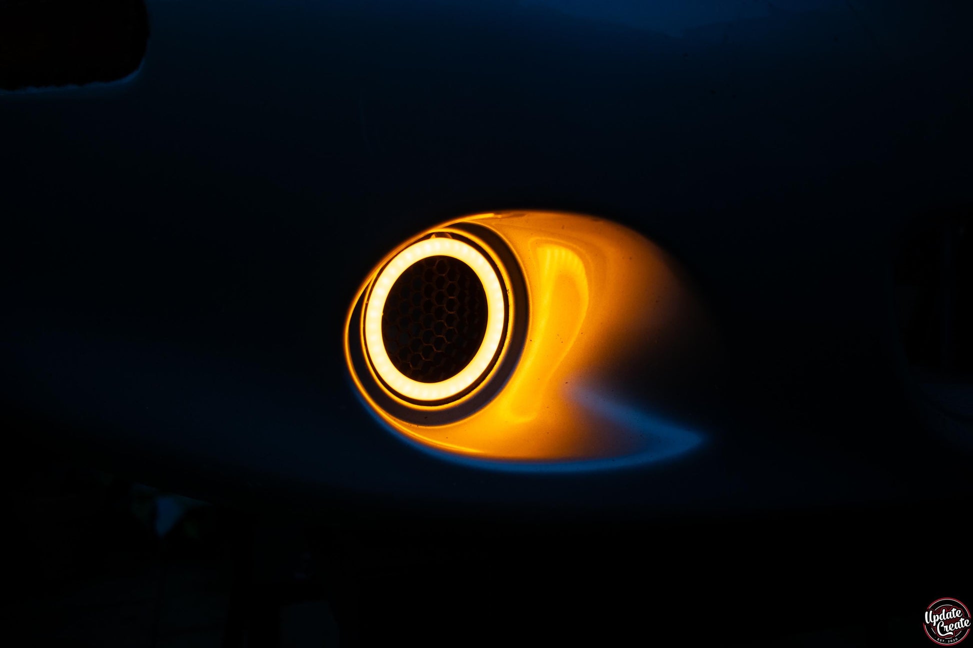 LED Halo light mounted to a custom 3D printed housing installed onto an OEM silver Mazda Mx5 NB bumper. The light is turned on, emitting an amber colour. The item is pictured at night to show its brightness in the dark.