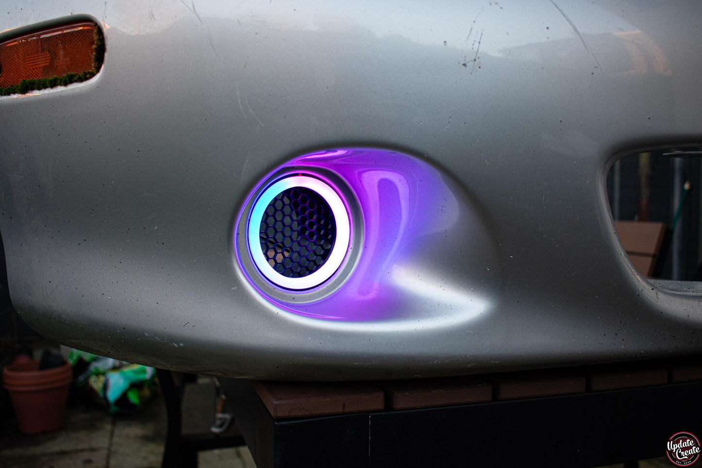RGB LED Halo light mounted to a custom 3D printed housing installed onto an OEM silver Mazda Mx5 NB bumper. The light is turned on, cycling through its RGB colours.