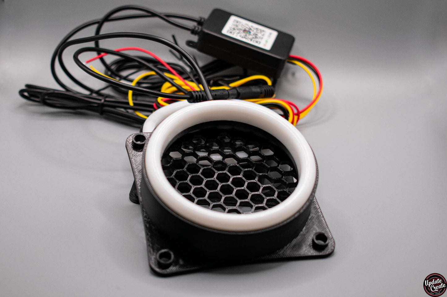 LED Halo light mounted to a custom 3D printed housing to fit the OEM fog light bumper opening on a Mk2 Mazda MX5/Miata. The item has a honeycomb pattern in the light opening to help reduce debris from being sent through it. Also pictured is the wiring and control box for the LED halos. Item is set in a well lit, light grey background.