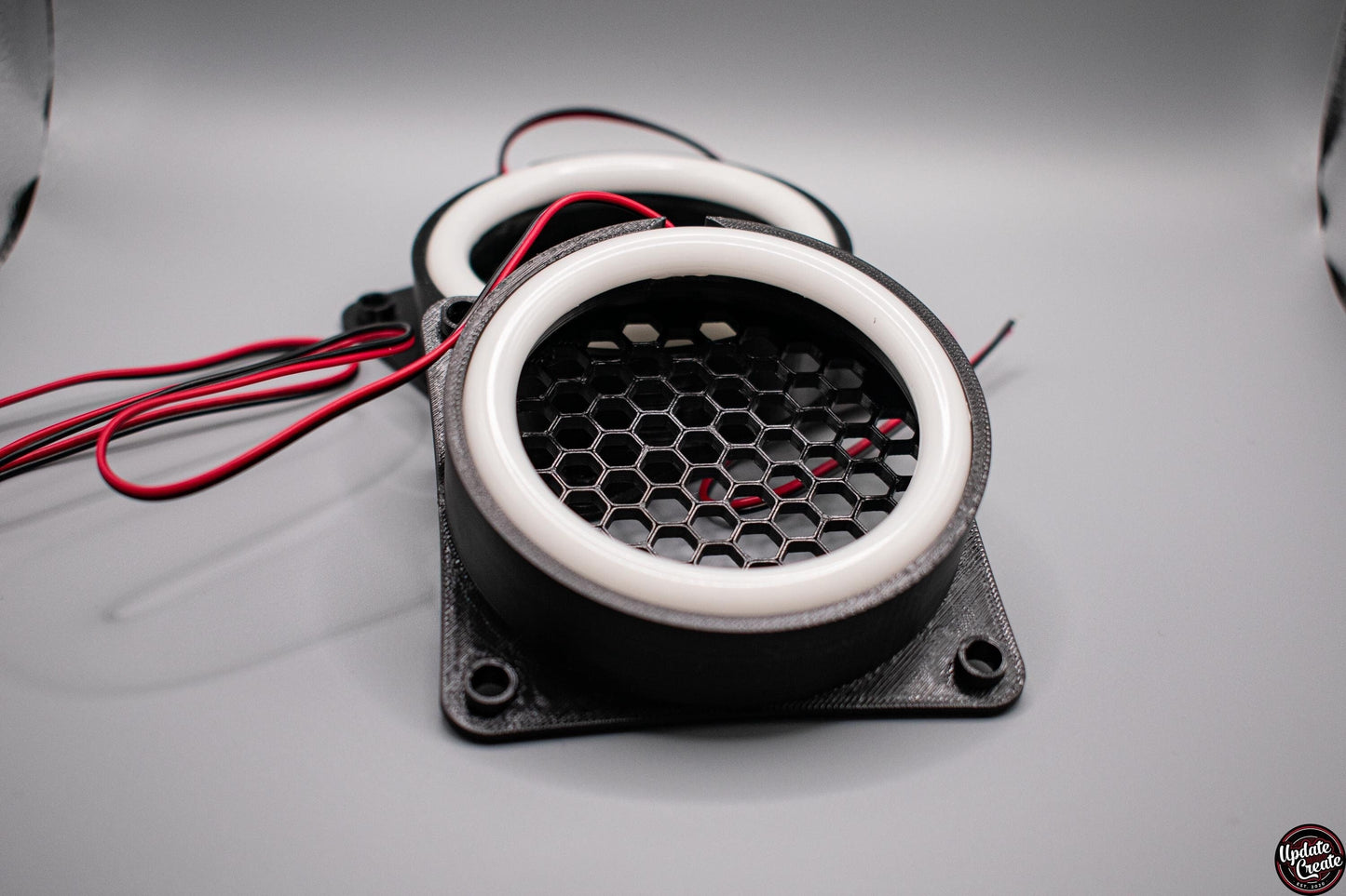 LED Halo light mounted to a custom 3D printed housing to fit the OEM fog light bumper opening on a Mk2 Mazda MX5/Miata. The item has a honeycomb pattern in the light opening to help reduce debris from being sent through it. Also pictured is the wiring and control box for the LED halos. Item is set in a well lit, light grey background.